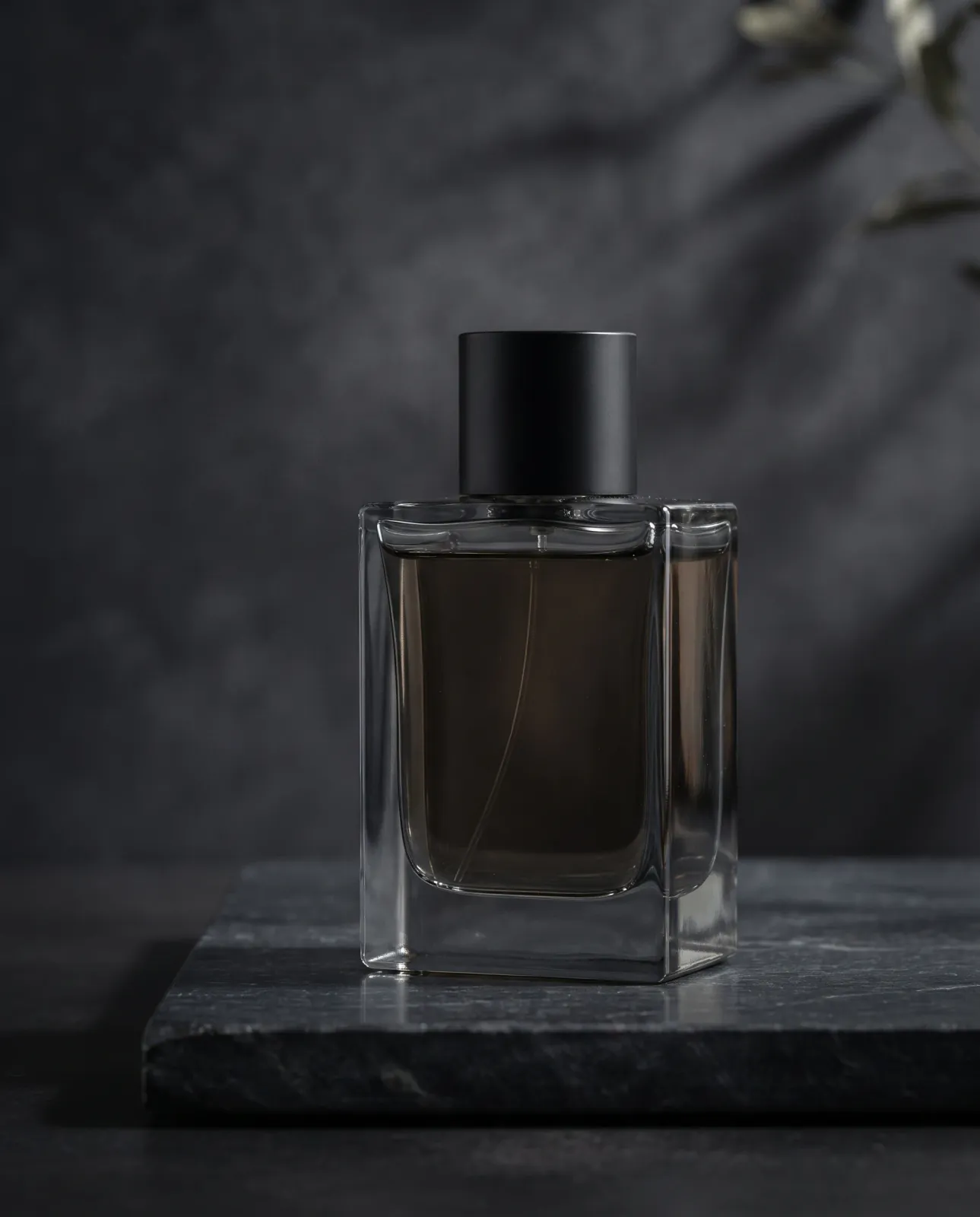 An unbranded rectangular glass perfume bottle with dark amber liquid and a matte-black cap in a smoky charcoal studio.