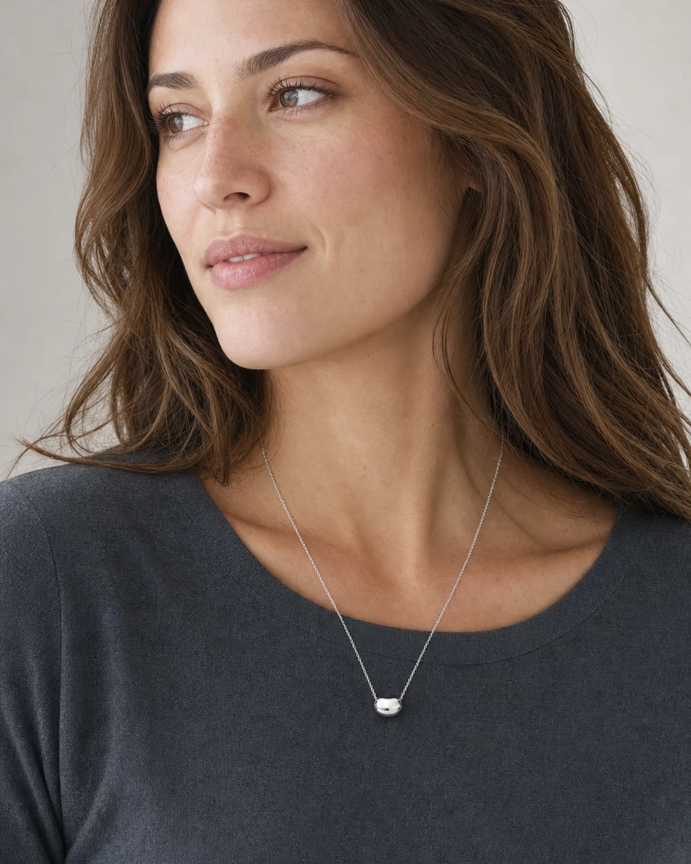 A woman wearing a Tiffany Elsa Peretti Bean pendant in sterling silver with the complete necklace clearly visible against a charcoal top.